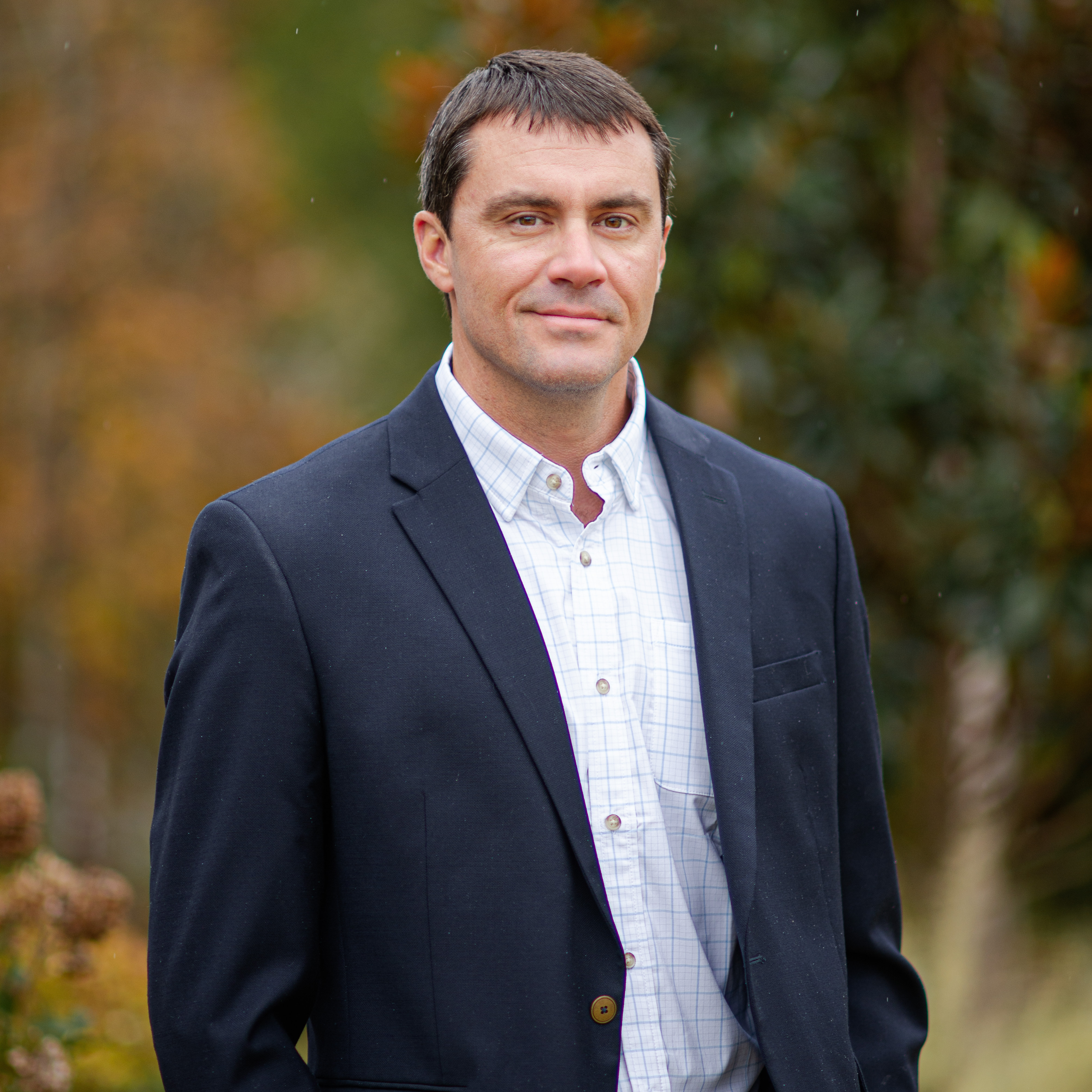 Professional portrait of Chad Lindlerwearing a navy blazer and white plaid shirt, standing outdoors with blurred greenery and autumnal tones in the background.