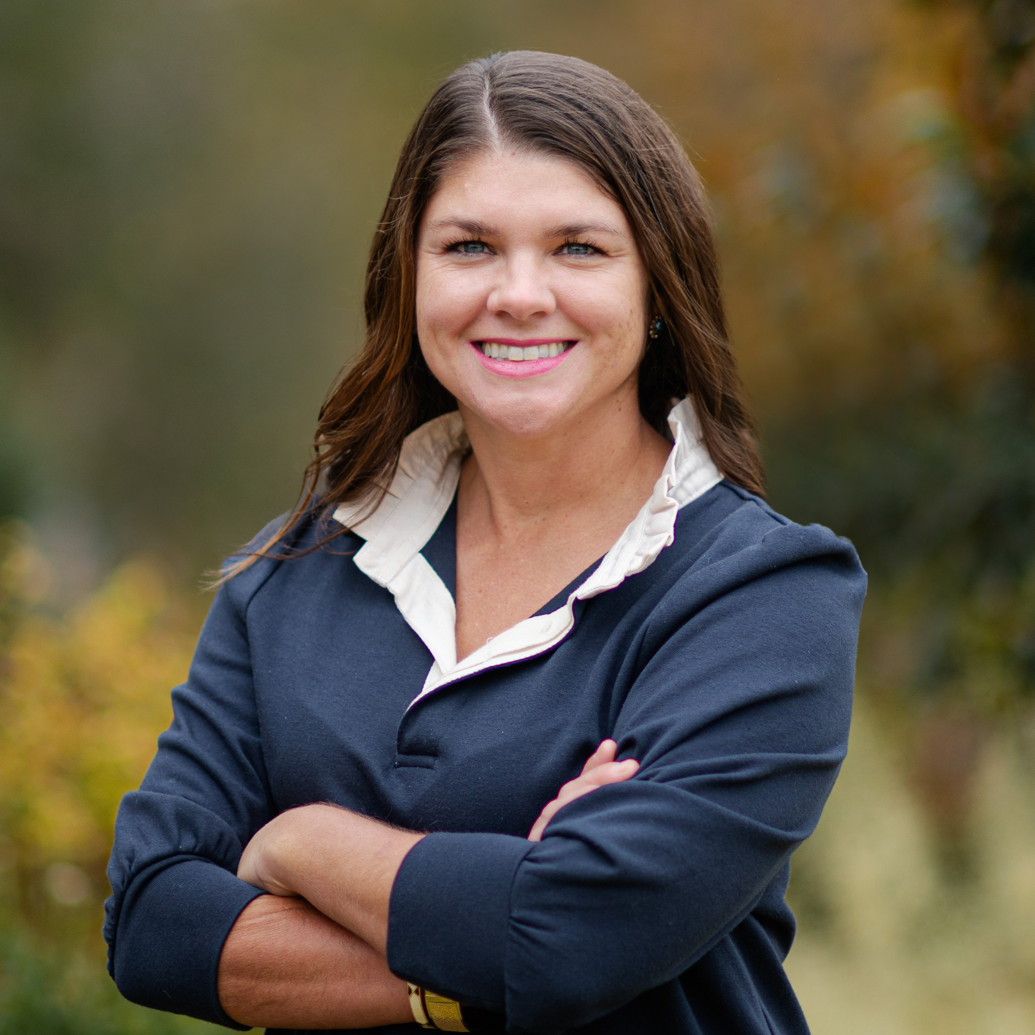 Professional portrait of Caroline Lindler, smiling with arms crossed, wearing a navy top with a white collar, standing outdoors against a blurred background of greenery and autumn tones.